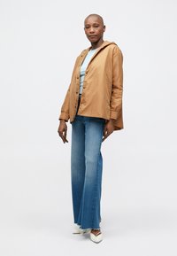 Hellbrauner, kapuzenloser Mantel mit Knopfverschluss, kombiniert mit einem hellblauen Oberteil und schmalen, blauen Jeans mit Farbverlauf. Weiße, spitze Schuhe runden das Outfit ab.