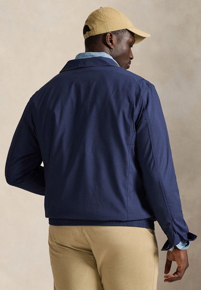 Mann mit beiger Kappe, dunkelblauer Jacke, hellblauem Hemd und beiger Hose, leicht abgewandt vor einem schlichten Hintergrund.