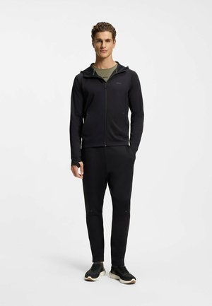 Schwarzer sportlicher Trainingsanzug mit Reißverschlussjacke, ausgestattet mit Kapuze und zwei Seitentaschen, kombiniert mit zulaufenden Hosen und eleganten schwarzen Sneakers.