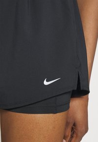 Černé sportovní šortky Nike mají hladkou texturu, rozdělený design na lemu a bílé logo na dolní levé straně.