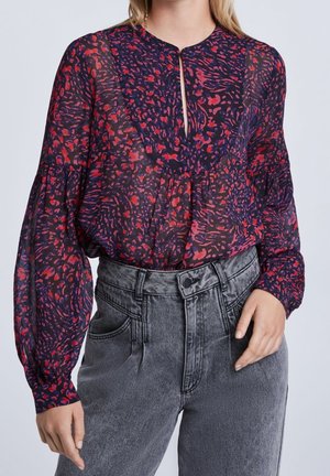 Femme portant un chemisier transparent à manches longues avec des motifs abstraits rouges et violets, rentré dans un jean noir délavé taille haute.