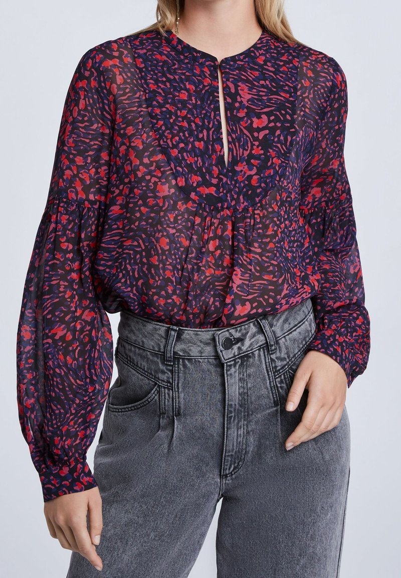 Femme portant un chemisier transparent à manches longues avec des motifs abstraits rouges et violets, rentré dans un jean noir délavé taille haute.