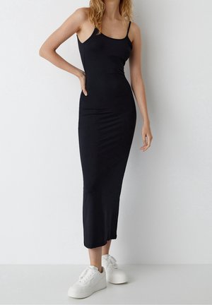 Femme portant une robe midi noire moulante sans manches et des baskets à plateforme blanches, debout avec une main sur la hanche contre un mur blanc uni.