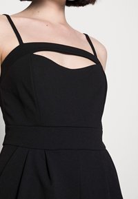 Svart ärmlös klänning med en åtsittande överdel, med ett utskärningsdetalj under halsringningen och en plisserad kjol. Slen, texturerad tyg.