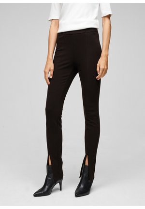 Pantalons noirs slim-fit avec des fentes sur les côtés au bas, fabriqués dans un tissu extensible. Portés avec des bottines noires à talons pointus, arborant une texture lisse.