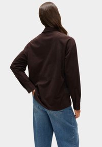 Hnědá košile s dlouhými rukávy a límcem, volného střihu s knoflíkovými manžetami, v kombinaci s světle modrými širokými denimovými džínami.