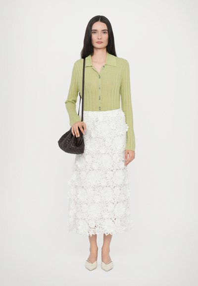 Lys grønn ribbestrikket cardigan med knappelukking, kombinert med et hvitt skjørt i blonder med blomster. Outfitten inkluderer en liten svart flettet håndveske og kremfargede hæler.