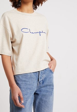 Osoba v béžovém tričku Champion a světle modrých džínách, s jednou rukou v kapse, na jednobarevném bílém pozadí.