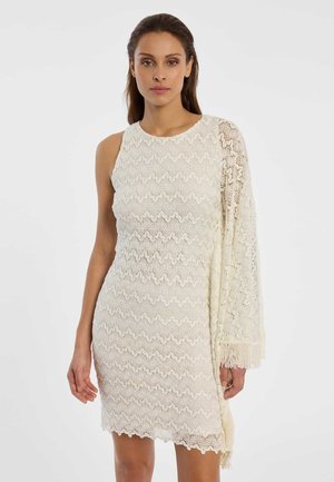 Femme portant une robe ajustée en dentelle blanche avec un côté sans manche et une manche longue en dentelle ornée de franges.