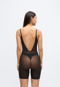 Fekete áttetsző body V-kivágással a háton, vékony pántokkal és a comb közepéig érő hosszúsággal. Az anyag könnyűnek és testhez simulónak tűnik, kiemelve a sziluettet.