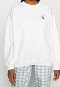 Sudadera blanca con cuello redondo, mangas globo y un pequeño diseño colorido de un colibrí en el área del pecho izquierdo.
