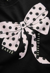 Didelė šviesiai rožinė dekoracija su juodais taškeliais, atspausdinta ant juodos medžiagos, su žodžiais "BYTED BAKER" ir "BAKER" balta spalva.