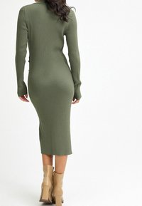 Robe à manches longues côtelée, verte olive, avec une silhouette ajustée et une longueur mi-mollet, assortie à des bottes à talons beiges.