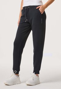 Femme portant un pantalon de jogging noir avec taille à cordon et des baskets blanches, une main dans la poche, debout sur un fond neutre.