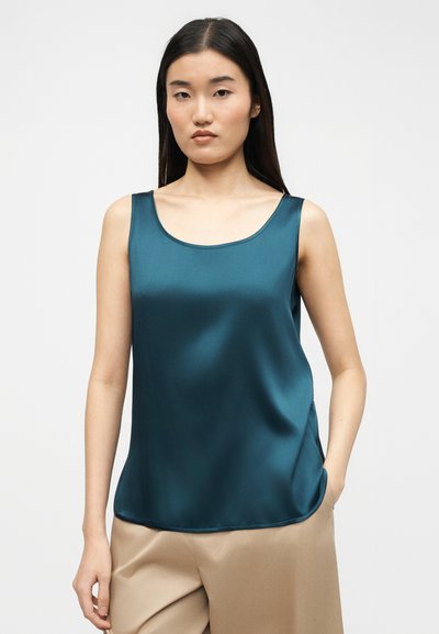 Γυναίκα με μακριά σκούρα μαλλιά, που φοράει αμάνικο σατέν τοπ σε τυρκουάζ χρώμα και μπεζ παντελόνι, στέκεται μπροστά σε απλό ανοιχτό φόντο.