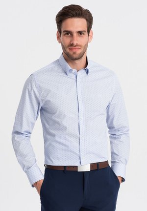 Lichtblauw overhemd met kleine donkerblauwe stippen, met puntkraag en lange mouwen. Draag met marineblauwe broek en een bruine riem.