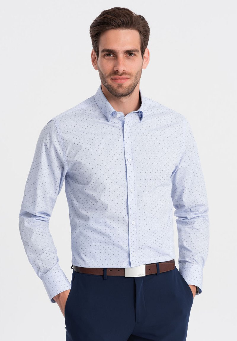 Camicia azzurra chiara con piccoli pois blu navy, colletto a punta e maniche lunghe. Indossata con pantaloni navy e una cintura marrone.