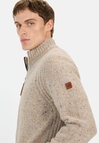 Beżowy sweter z dzianiny z żeberkowym kołnierzem, zamkiem przy szyi oraz fakturowanym materiałem z niebieskimi i pomarańczowymi plamkami. Skórzana naszywka z logo na rękawie.