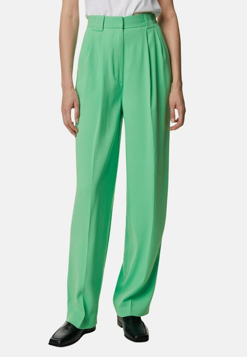 Personne portant un pantalon vert taille haute à jambes larges, des chaussures noires à bouts carrés et une chemise blanche à manches courtes rentrée, debout devant un fond blanc.