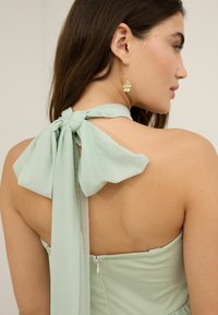 Mintgrön klänning med halterneck, knuten i ryggen med en svävande rosett. Har en dragkedjeslutning och en mjuk tygtextur.