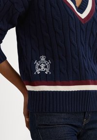 Laivaston sininen palmikkoneulepaita V-aukolla, jossa on viininpunainen ja valkoinen raidallinen nauha helman kohdalla sekä kirjailtu logoyksityiskohta rinnassa.