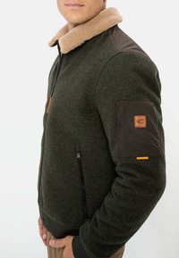 Fleece-lined jacket in dark green with brown leather accents. Features a tan faux fur collar and a zippered front with side pockets.