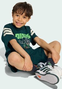 Zelené bavlněné tričko s bílými pruhy, které má potisk s postavou. Sestaveno s odpovídajícími kraťasy a teniskami s zelenými akcenty.