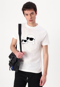White cotton t-shirt featuring a black graphic of a cat and a figure in sunglasses. Model holds a black crossbody bag with multiple pockets.