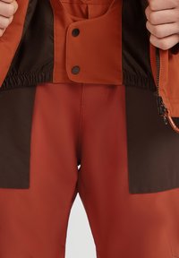 Waterproof oranje jack met bruine accenten, voorzien van een drukknoopsluiting, elastische taille en een ritszak. Gelaagd stof met een gladde voering.