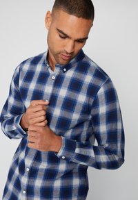 Blå og hvid ternet skjorte lavet af flannel, med button-down krave, lange ærmer og knapmanchetter. Fremhævet med lyseblå firkanter.