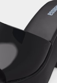 Μαύρο γυαλιστερό σανδάλι πλατφόρμας με ανοιχτά δάχτυλα, χοντρή σόλα και το όνομα της μάρκας τυπωμένο στη σόλα του ποδιού.