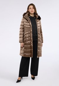 Abrigo largo acolchado marrón con textura brillante, capucha con borde de piel y cremallera frontal. Combinado con una camiseta oscura y pantalones anchos negros.