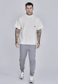 Biele bavlnené tričko s krátkym rukávom a čiernym logom, v kombinácii so sivými teplákmi a bielymi športovými topánkami. Jednoduchý, ležérny dizajn.
