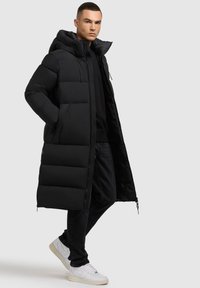 Abrigo acolchado negro con capucha, que presenta un diseño largo y acolchado, cierre de cremallera y dos bolsillos laterales. Combinado con jeans negros y zapatillas blancas.