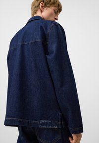 Mörkblå denimjacka med klassisk skärning, synliga sömmar och en etikett vid fållen. Strukturerad tyg med en något blekt utseende.