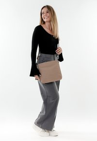 Junge Frau trägt ein schwarzes Langarmshirt, graue Hosen mit weitem Bein, weiße Turnschuhe, trägt eine beige Schultertasche, lächelt und schaut zur Seite.