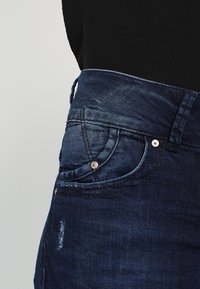Jeans in denim blu scuro con una vita alta, dettagli sulle tasche frontali, leggero sbiadimento e un accento in pulsante argentato. La texture è liscia.