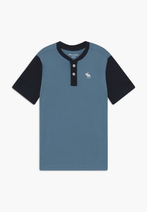Koszulka henley z krótkim rękawem, niebieskim korpusem, czarnymi rękawami i kołnierzem, dwoma przednimi guzikami oraz małym białym logo łosia na piersi.