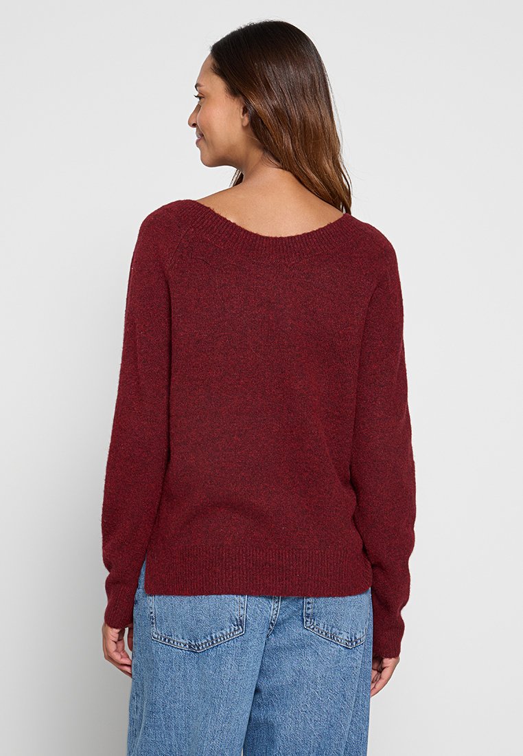Pull en tricot bordeaux avec un col large et des manches longues. Texture douce, poignets et ourlet côtelés, associé à un jean bleu à coupe ample.