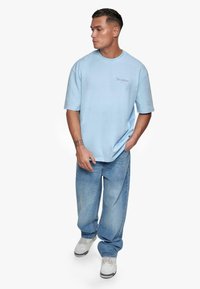 Lyseblå kortermet t-skjorte med løs passform og brodert tekst på brystet, kombinert med avslappet blå jeans og grå joggesko.