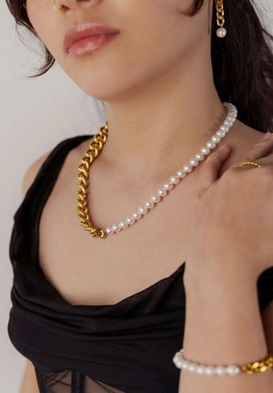 Woman wearing mixed gold chain and pearl necklace, matching pearl and gold bracelet, pearl earring, nose ring, and gold ring with black top.