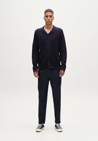 Marinblå stickad cardigan med kragkrage och knappstängning, tillsammans med mörk denimbyxor. Modellen har på sig svart-vita sneakers.