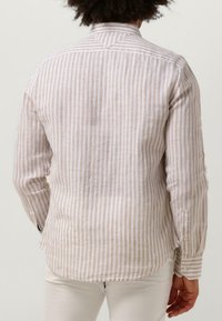Man in een beige en witte verticaal gestreepte lange mouwen shirt en witte broek, van achteren weergegeven tegen een effen achtergrond.