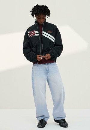 Persoană cu dreadlocks care poartă o jachetă neagră cu fermoar și dungi albe, blugi largi de un albastru deschis și adidași negri, stând pe un fundal simplu.