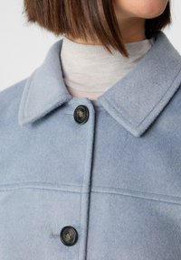 Jasnoniebieska kurtka z mieszanki wełny z odwróconym kołnierzem i zapięciem na guziki z przodu z czarnym detalem guzika. Miękka tekstura.
