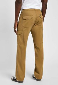 Pantaloni cargo beige con più tasche posteriori, materiale resistente, gambe dritto e dettagli di cucitura visibili; indossati con sneakers nere e bianche.