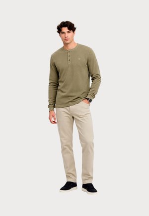 Mand iført olivengrøn langærmet henley-trøje, beige bukser, sorte sneakers og ur, står med den ene hånd i lommen mod en hvid baggrund.