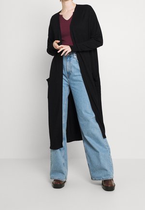 Femme portant un long cardigan noir ouvert, un haut bordeaux à col en V, un jean large taille haute bleu clair, et des chaussures noires, debout avec les mains près de la taille.