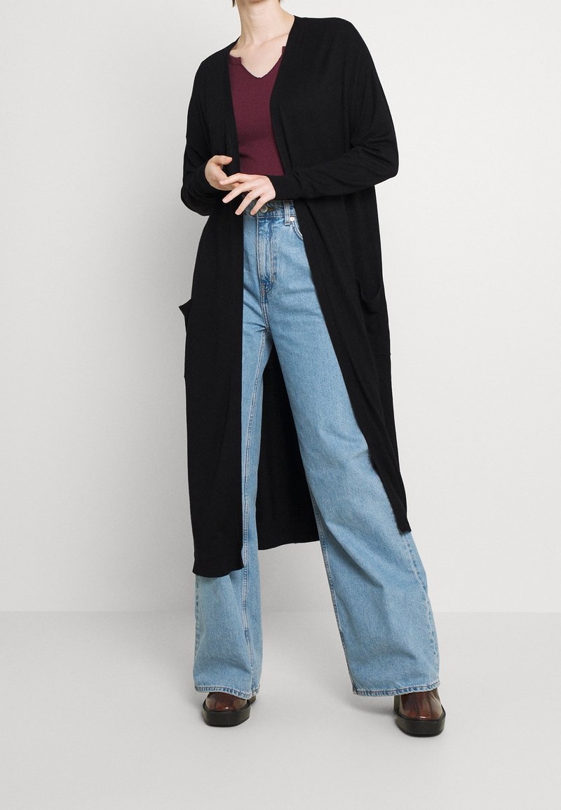 Femme portant un long cardigan noir ouvert, un haut bordeaux à col en V, un jean large taille haute bleu clair, et des chaussures noires, debout avec les mains près de la taille.