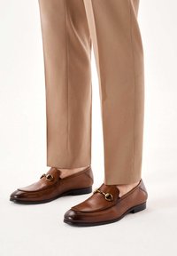 Mocasines de cuero marrón con un acabado pulido, que cuentan con un detalle decorativo en color dorado y un tacón bajo, combinados con pantalones de corte ajustado color beige.